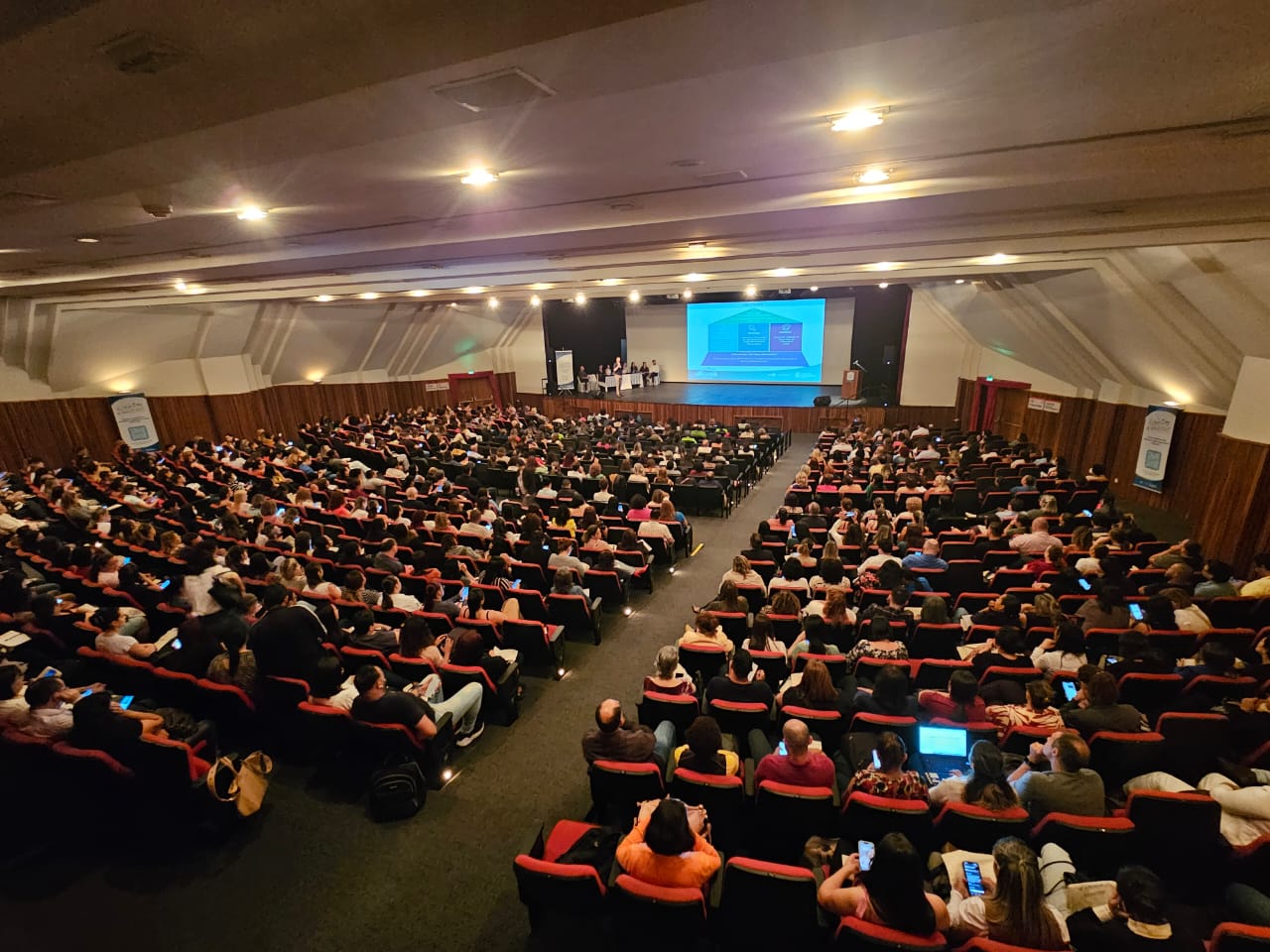 A foto mostra a visão completa da plateia de um auditório, vista de trás; ao fundo está o palco, a imagem de um telão